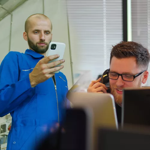 Auf diesem Foto sind zwei Männer abgebildet. Ein Mann guckt in das Smartphone in seiner Hand. Der andere sitzt und hat ein Telefonhörer an seinem Ohr.